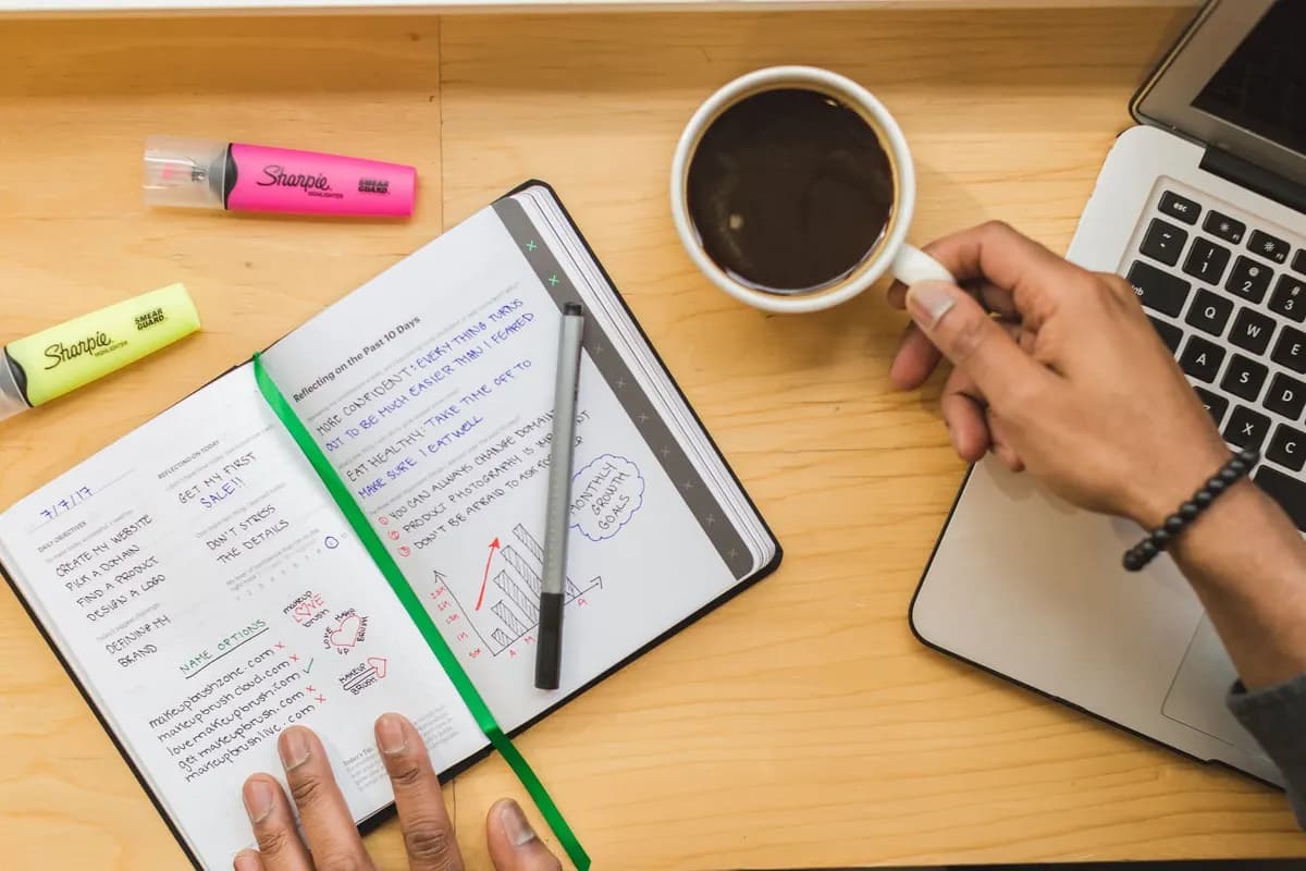 A notebook, coffee and laptop on a wooden desk, seen from above